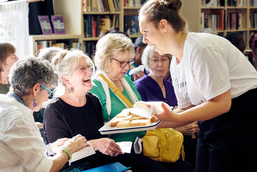 A young white woman, a cast member of the show show on Suffragettes is offering and older, female audience member a slice of cake. They are both laughing