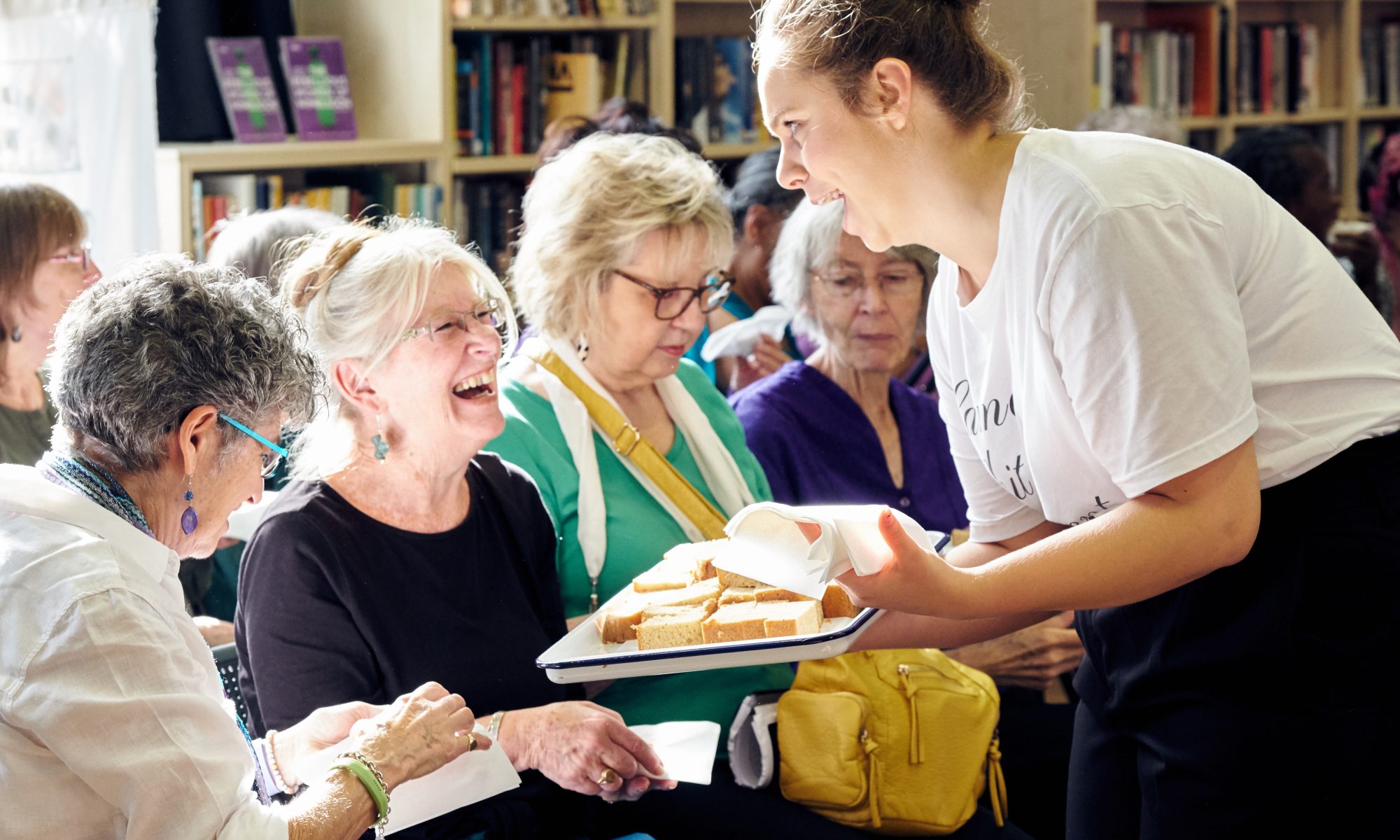 A young white woman, a cast member of the show show on Suffragettes is offering and older, female audience member a slice of cake. They are both laughing